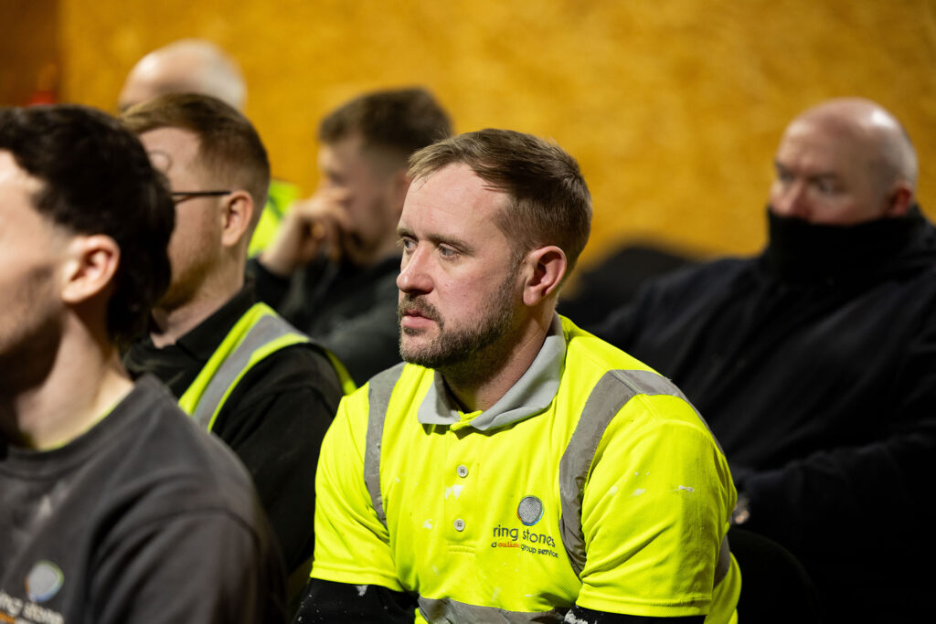 Adult learner in high‑visibility workwear seated during a live training session, listening to course delivery