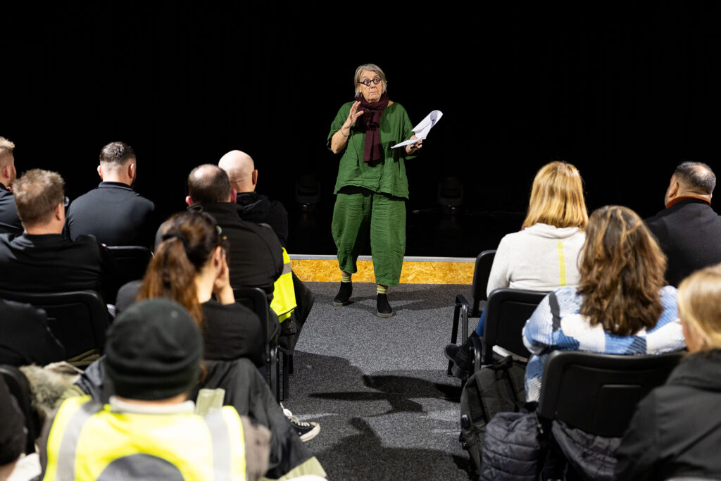 Tutor standing at the front of a live training session, holding notes and addressing seated adult learners.